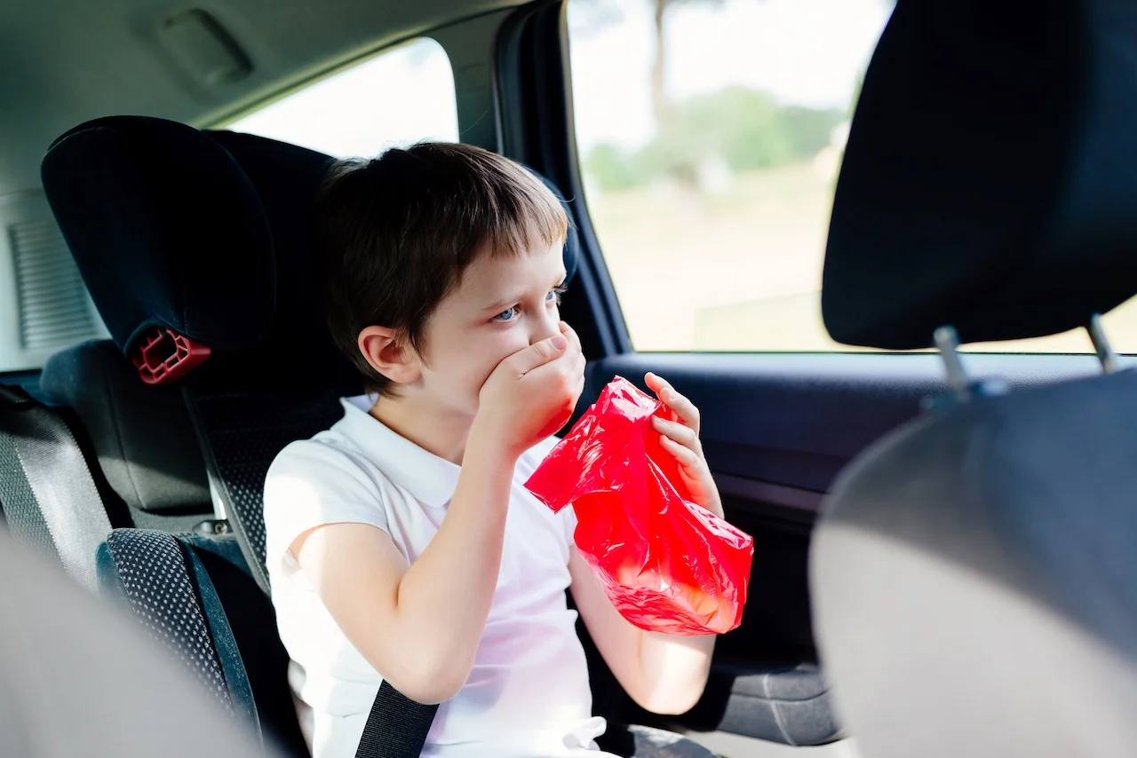 KIlkuletni chłopiec siedzi w foteliku samochodowym i wymiotuje do czerwonej torebki z powodu choroby lokomocyjnej